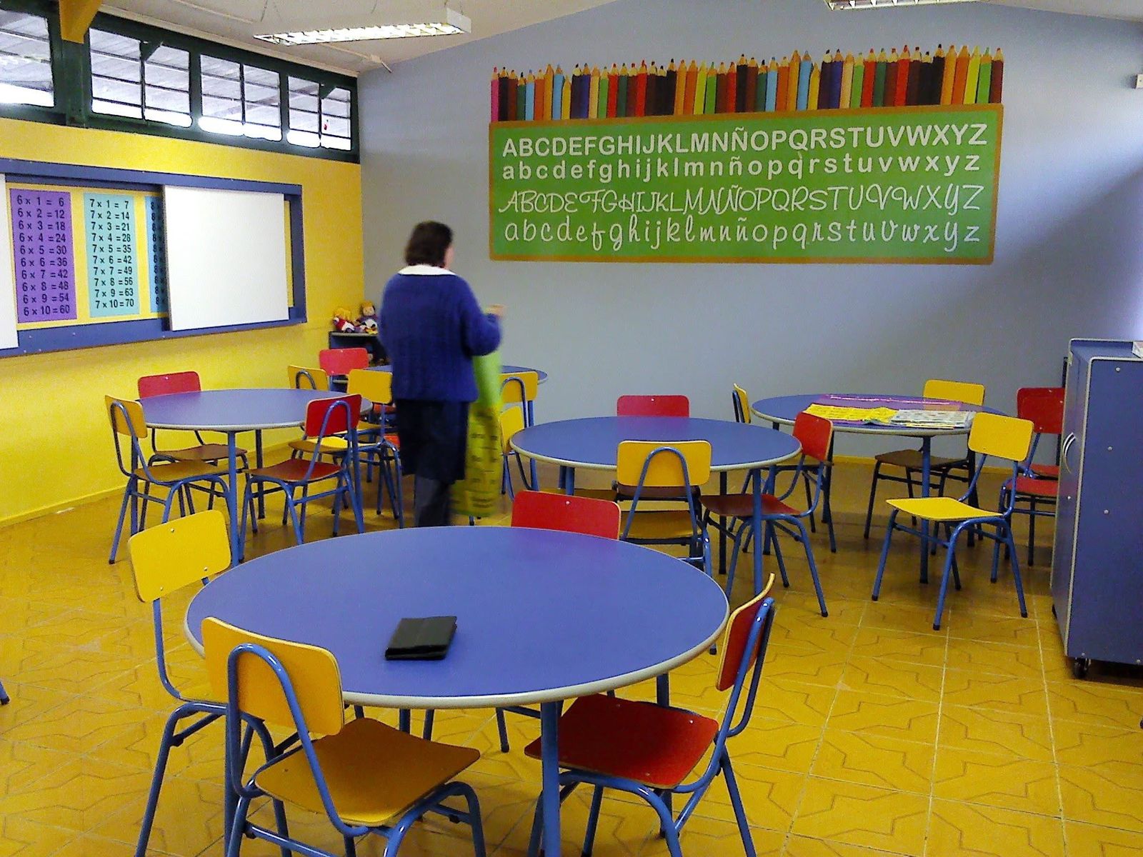 Aula de lenguaje con mesas redondas azules, sillas de colores y mural del abecedario